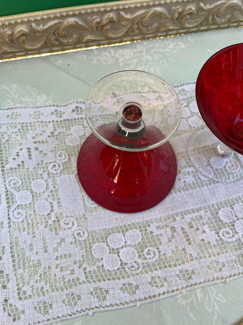 May include: Two vintage red glass goblets with clear stems and bases. The goblets are set on a white lace tablecloth. The goblets have a rounded bowl shape. The background includes a decorative gold-framed mirror.