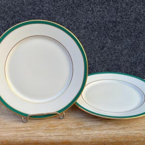 May include: Two round white ceramic plates featuring a wide dark green border along the rim, accented by a thin gold line. One plate stands upright on a metal display stand, while the second plate lies flat beside it on a wooden surface.