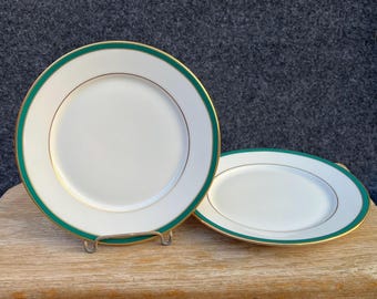 May include: Two round white ceramic plates featuring a wide dark green border along the rim, accented by a thin gold line. One plate stands upright on a metal display stand, while the second plate lies flat beside it on a wooden surface.