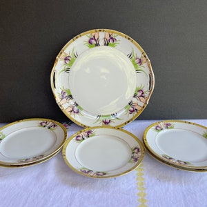 May include: Set of six vintage porcelain plates with a floral design. The plates feature a white background with purple and green flowers and gold accents. The set includes a large serving platter and five smaller plates.