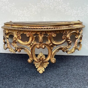 May include: A semi-circular wall-mounted console shelf featuring an ornate gold finish. The structure displays intricate carved scrollwork, acanthus leaf motifs, and a decorative central pendant element extending downward from the curved frame.