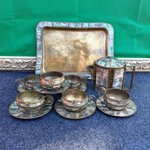 May include: A vintage tea set with six cups and saucers, a teapot, and a serving tray. The set features a silver-toned metal with decorative mosaic accents in shades of blue, green, and orange. The cups have ornate handles.
