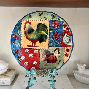May include: A decorative ceramic plate featuring a rooster design. The plate has a blue, green, red, and yellow color scheme. The plate is supported by a bronze-colored metal stand with a floral and leaf design.