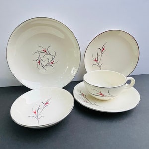 May include: A cream-colored vintage dish set with black and red floral designs. The set includes a large plate, a smaller plate, a bowl, a teacup, and a saucer. Each piece has a silver trim.