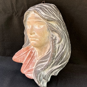 May include: A plaster sculpture of a woman's head and shoulders. The sculpture is painted in shades of brown, pink, and white. The woman has long, dark hair and is wearing a necklace.