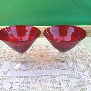 May include: Two ruby red martini glasses with clear stems and bases. The glasses are set on a white lace tablecloth. The background features a green surface and a decorative gold frame.