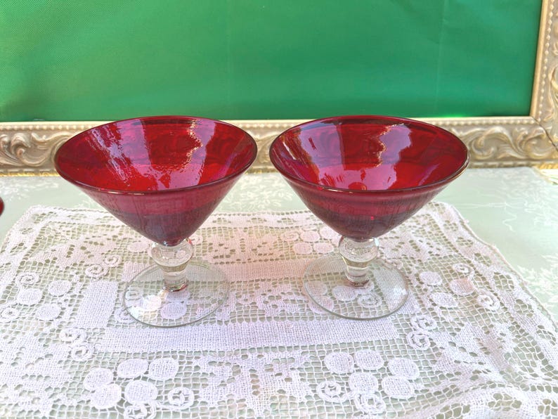 May include: Two ruby red martini glasses with clear stems and bases. The glasses are set on a white lace tablecloth. The background features a green surface and a decorative gold frame.