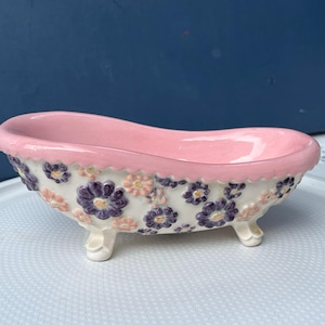 May include: A small, decorative ceramic bathtub with a pink interior and rim. The exterior is white, adorned with purple and peach-colored floral designs. The tub rests on three small, white feet.