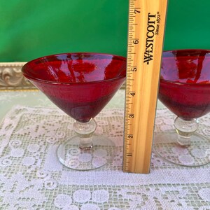 May include: Two red cocktail glasses with clear stems and bases. A wooden ruler is placed next to the glasses. The glasses are on a white lace tablecloth. The background is green.