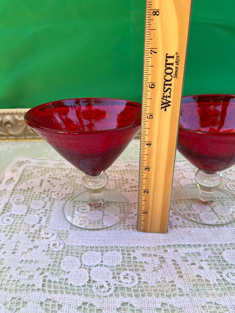 May include: Two red cocktail glasses with clear stems and bases. A wooden ruler is placed next to the glasses. The glasses are on a white lace tablecloth. The background is green.