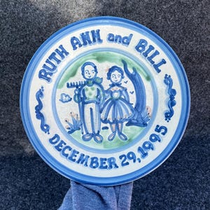 May include: A decorative ceramic plate with a blue and white border. The plate features a hand-painted illustration of a couple and the text "RUTH ANN and BILL" and "DECEMBER 29,1995". The plate has a vintage, folk art style.