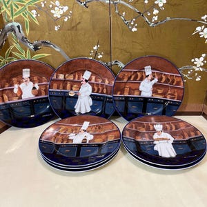May include: Set of six blue and brown ceramic dinner plates with a chef theme. Each plate features a different chef in a white uniform and a blue and brown kitchen scene.