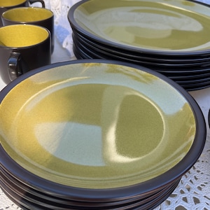 May include: A stack of olive green plates with dark brown rims. Several matching coffee mugs with olive green interiors and dark brown exteriors are also visible. The plates and mugs are arranged on a white lace tablecloth.