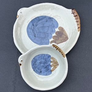 May include: Two ceramic plates with a bird design. The plates are white with brown and blue accents. The larger plate has a brown rim and the smaller plate has a brown handle.