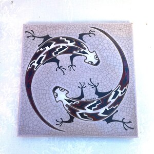 May include: A square ceramic tile with a light beige crackle glaze finish featuring two stylized lizards arranged in a circular pattern. The lizards have black, white, and dark red geometric markings on their bodies and the name Tongier is printed.
