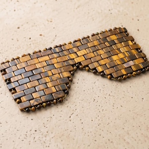 May include: A sleep mask made of rectangular, tiger's eye stones. The mask is a mosaic of brown, tan, and gold hues, with small, round beads along the edges. The mask is designed to cover the eyes for light blocking.