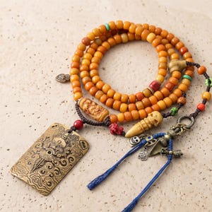 Op de afbeelding: Een sierlijke ketting met amberkleurige kralen, decoratieve hangers en een rechthoekige messing bedel met ingewikkelde ontwerpen. De ketting bevat een verscheidenheid aan kralen in verschillende kleuren en vormen, en blauwe kwastjes.