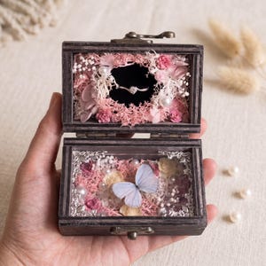 May include: A dark wooden box with a hinged lid, featuring a silver ring nestled in pink preserved flowers and white pearls in the top section, and a light blue butterfly surrounded by pink and cream flowers in the bottom section.