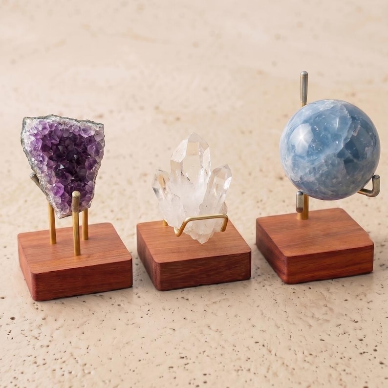 Op de afbeelding: Drie kristallen displays. De eerste toont een paarse amethistcluster, de tweede een heldere bergkristal en de derde een blauwe bol. Elk is gemonteerd op een kleine, vierkante houten basis met metalen steunen.