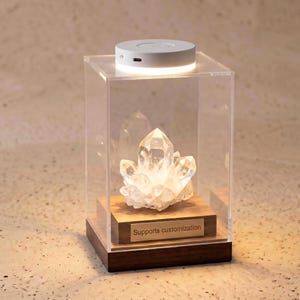 May include: A clear quartz crystal cluster displayed inside a clear acrylic box with a wooden base and a small light on top. The wooden base has a small plaque that reads "Supports customization".