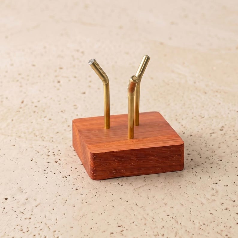 Op de afbeelding: Een vierkant, roodbruin houten blok met drie messingkleurige metalen stelen. De stelen zijn aan de bovenkant gebogen. Het object is waarschijnlijk een decoratief item of een houder voor kleine items.