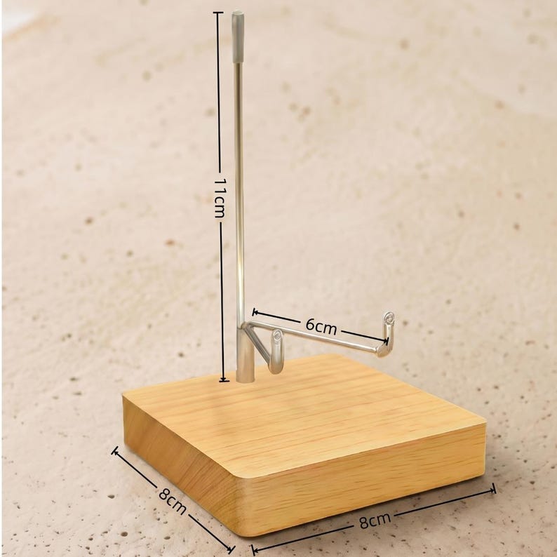 Op de afbeelding: Een houten telefoonstandaard met een zilverkleurige metalen houder. De basis is een lichtbruin vierkant, 8 cm bij 8 cm. De metalen houder is 11 cm hoog en 6 cm breed. De standaard is ontworpen om een telefoon vast te houden.