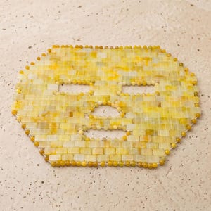 Op de afbeelding: Een gele jade gezichtsmasker met uitsparingen voor ogen, neus en mond. Het masker is gemaakt van kleine, rechthoekige stukjes jade en ronde kralen, verbonden door witte draad. Het masker is ontworpen om het gezicht te bedekken.