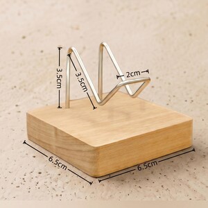 Op de afbeelding: Een houten displaystandaard met een lichtbruine afwerking en een metalen draadframe. De standaard meet 6,5 cm x 6,5 cm aan de basis, met het draadframe dat 3,5 cm hoog en 2 cm breed is. Geschikt voor het presenteren van kleine items.