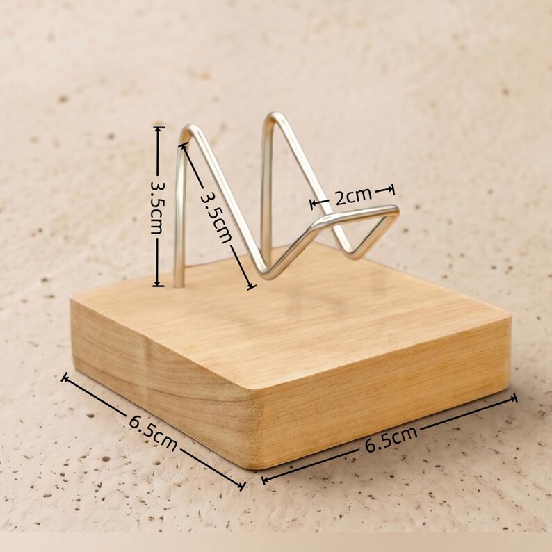 Op de afbeelding: Een houten displaystandaard met een lichtbruine afwerking en een metalen draadframe. De standaard meet 6,5 cm x 6,5 cm aan de basis, met het draadframe dat 3,5 cm hoog en 2 cm breed is. Geschikt voor het presenteren van kleine items.