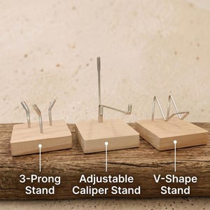 Op de afbeelding: Drie houten display stands met metalen steunen. De stands zijn gelabeld: "3-Prong Stand", "Adjustable Caliper Stand" en "V-Shape Stand". De stands staan op een houten oppervlak.