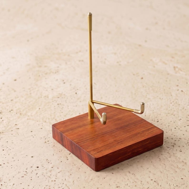 Op de afbeelding: Een displaystandaard met een vierkante, roodbruine houten basis. Een goudkleurige metalen steun komt omhoog vanaf de basis, met een horizontale arm en twee kleine haken. De standaard is ontworpen om een kleine plaat of ander decoratief item te bevatten.