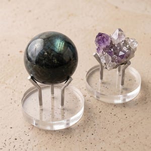 Puede incluir: Dos exhibidores decorativos de cristales. Uno presenta una esfera oscura e iridiscente, y el otro muestra un grupo de cristales morados y transparentes. Ambos están montados sobre bases de acrílico transparente con soportes plateados.