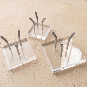 May include: Three clear acrylic display stands with silver metal posts. Each stand has a square base and multiple posts angled upwards. The stands are arranged on a light-colored, textured surface.