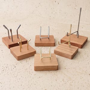 Op de afbeelding: Een verzameling kleine display stands. Elke stand heeft een vierkante houten basis met een metalen steun in verschillende vormen en afwerkingen, waaronder zilver en goud. De stands zijn ontworpen om kleine items vast te houden.