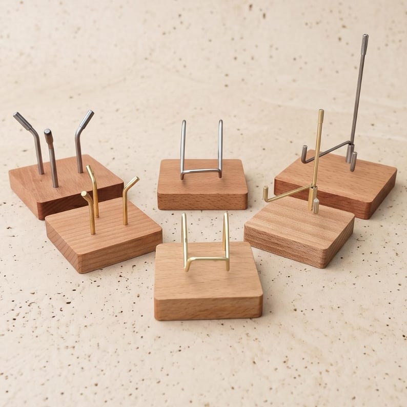 Op de afbeelding: Een verzameling kleine display stands. Elke stand heeft een vierkante houten basis met een metalen steun in verschillende vormen en afwerkingen, waaronder zilver en goud. De stands zijn ontworpen om kleine items vast te houden.
