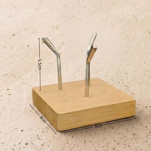 Op de afbeelding: Een houten displaystandaard met twee zilveren metalen haken. De basis is een lichtgekleurde vierkant, 8 cm bij 8 cm. De haken zijn 5,5 cm hoog. De standaard is ontworpen om kleine items vast te houden.
