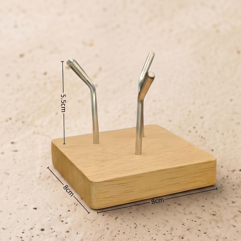Op de afbeelding: Een houten displaystandaard met twee zilveren metalen haken. De basis is een lichtgekleurde vierkant, 8 cm bij 8 cm. De haken zijn 5,5 cm hoog. De standaard is ontworpen om kleine items vast te houden.