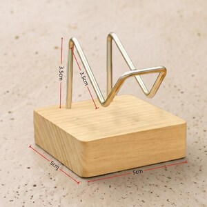 Op de afbeelding: Een houten telefoonstandaard met een zilverkleurige metalen houder. De houten basis is een lichtbruin vierkant, 5 cm aan elke kant. De metalen houder is 3,5 cm hoog en heeft een driehoekige vorm.