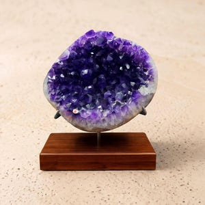 Op de afbeelding: Een grote amethistgeode met dieppaarse kristallen, gemonteerd op een donkerbruine houten basis. Dit decoratieve stuk toont de natuurlijke schoonheid van de edelsteen. De kristallen hebben een rijke, levendige paarse kleur.