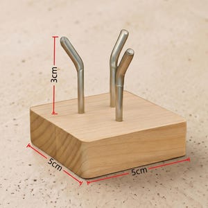 Op de afbeelding: Een houten blok met drie zilveren metalen stelen. Het blok is 5 cm x 5 cm en 3 cm hoog. De stelen zijn gebogen en lijken bedoeld om iets vast te houden. Het hout is licht van kleur en heeft een natuurlijke nerf.