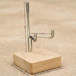 Op de afbeelding: Een displaystandaard met een houten basis van 5 cm bij 5 cm. Een zilveren metalen arm strekt zich 7 cm omhoog uit, met een gebogen haak van 3,5 cm. De standaard is ontworpen om een item vast te houden.