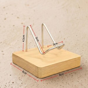 Op de afbeelding: Een houten telefoonstandaard met een zilveren metalen houder. De houten basis is 8 cm x 8 cm. De metalen houder is 5 cm hoog en 3 cm breed. De standaard is 4,5 cm hoog.