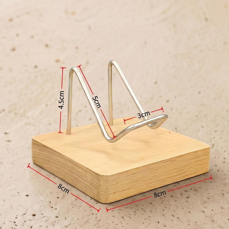 Op de afbeelding: Een houten telefoonstandaard met een zilveren metalen houder. De houten basis is 8 cm x 8 cm. De metalen houder is 5 cm hoog en 3 cm breed. De standaard is 4,5 cm hoog.
