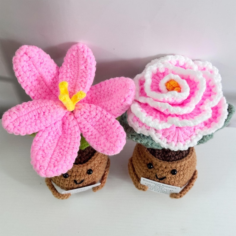 May include: Two crocheted flower pot figurines. The first is a pink flower with a yellow center in a brown pot with a smiling face. The second is a white and pink flower with a green base in a brown pot with a smiling face.