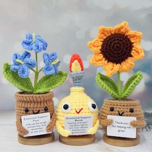 May include: Three crocheted plush toys shaped like plants in pots. The first is a blue flower with a sign that reads "Emotional Support Plant. Life can be tough sometimes, but remember so are you!" The second is a yellow bomb with a sign that reads "Emotional Support Bomb. Feeling stressed? Squeeze tightly, aim and release. Satisfaction guaranteed!" The third is a sunflower with a sign that reads "Thank you for helping me grow."