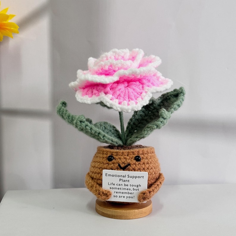 May include: A crocheted plant with a pink flower and green leaves. The plant is in a brown pot with a smiling face and is holding a sign that says "Emotional Support Plant. Life can be tough sometimes, but remember so are you!"