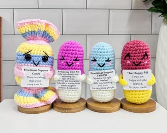 May include: Four crocheted pill-shaped plushies with happy faces and messages on cards. The plushies are in the colours pink, blue, yellow, and rainbow. The messages on the cards are "Emotional Support Candy", "Emergency Chill Pill", "Emotional Support Chill Pill", and "The Happy Pill".