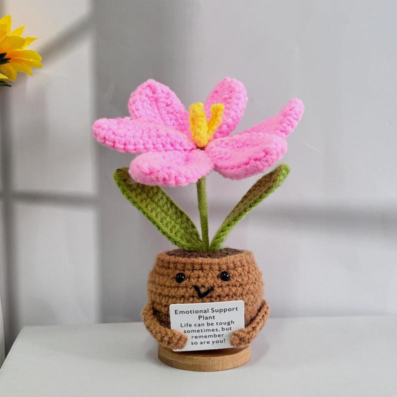 May include: A crocheted pink flower with green leaves in a brown pot with a smiling face. The pot is holding a card that reads "Emotional Support Plant Life can be tough sometimes, but remember so are you!"
