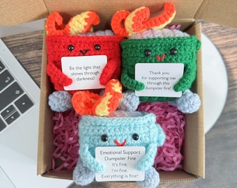Peut inclure: Trois jouets en peluche crochetés dans une boîte cadeau. Chaque jouet a la forme d'un feu avec un visage et un message. Les messages sont "Soyez la lumière qui brille dans l'obscurité !" "Merci de me soutenir dans ce brasier !" et "Feu de décharge émotionnel C'est bon. Je vais bien. Tout va bien !"