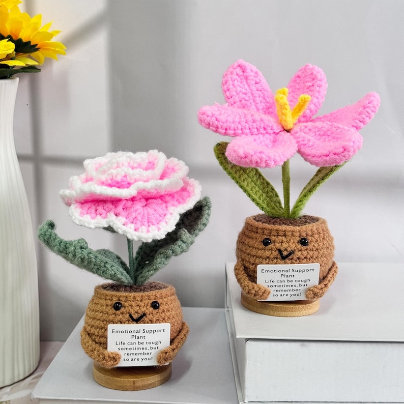 May include: Two crocheted flower plants in brown pots with smiling faces. Each pot has a small sign that reads "Emotional Support Plant" and "Life can be tough sometimes, but remember so are you!"